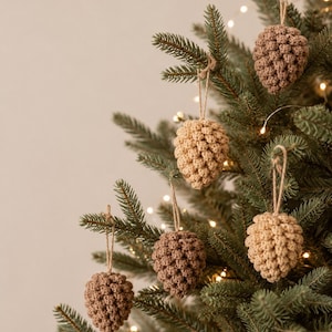 May include: Five crocheted pinecone ornaments in varying shades of tan and brown. Each ornament features a textured, knotted surface and is attached to a thin, light brown twine loop for hanging. The pinecones are displayed hanging from green branches.