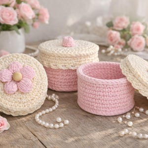 May include: Three handmade crochet boxes in cream and pink hues. One box features a pink flower, another has a cream lid with a pink trim, and the third is open. Pearls and pink roses decorate the wooden surface.