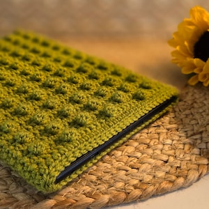 May include: A green crocheted cover for a tablet or e-reader. The cover has a textured, bumpy design and is lying on a woven straw mat.