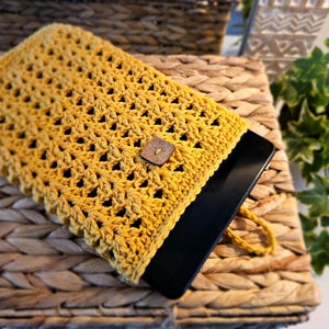May include: Mustard yellow crocheted tablet sleeve with a square wooden button fastening. The sleeve has an openwork design and a small loop for carrying. The tablet is black and partially visible.