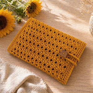 May include: Mustard yellow crochet tablet sleeve with a button closure. The sleeve has an openwork design and a rectangular shape. The background includes sunflowers, a vase with dried flowers, and a beige cloth on a wooden surface.