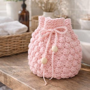 Puede incluir: Una bolsa con cordón rosa claro, tejida a ganchillo, con un patrón texturizado en forma de burbujas. La bolsa tiene un cierre de cordón rosa a juego con dos cuentas de madera. La bolsa está sobre una superficie de madera.