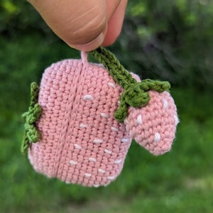 May include: A handmade crochet strawberry in pink and green. The strawberry features white speckles and a green stem. The crochet work is detailed, showing the yarn's texture. The strawberry is held against a blurred green background.