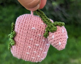 Pode incluir: Um morango de crochê feito à mão em tons de rosa e verde. O morango tem salpicos brancos e um caule verde. O trabalho de crochê é detalhado, mostrando a textura da linha. O morango é segurado contra um fundo verde desfocado.