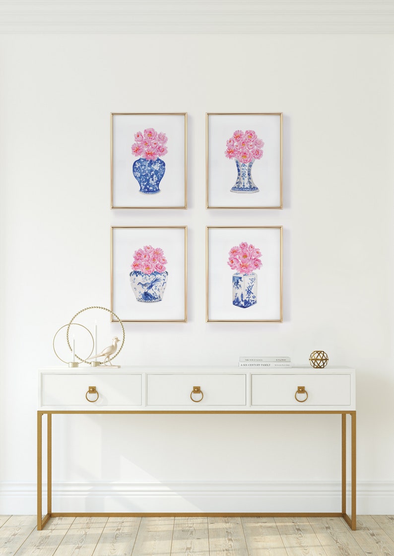 May include: Four framed prints of pink flowers in blue and white porcelain vases. The prints are arranged in a grid pattern on a white wall above a white console table with gold accents.
