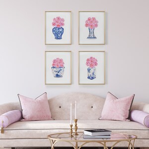 May include: Four framed prints of pink flowers in blue and white porcelain vases. The prints are arranged in a grid on a light pink wall above a white sofa with pink pillows.