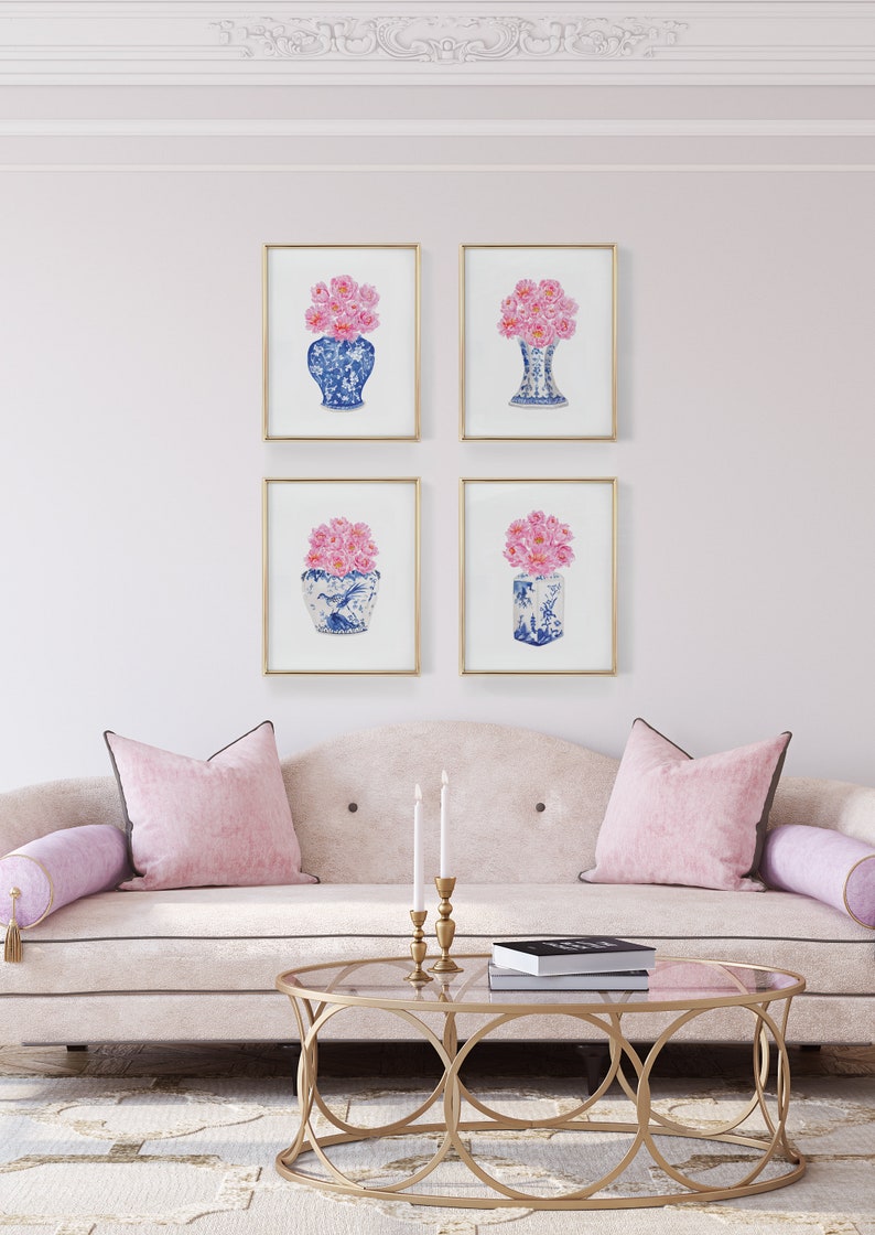 May include: Four framed prints of pink flowers in blue and white porcelain vases. The prints are arranged in a grid on a light pink wall above a white sofa with pink pillows.