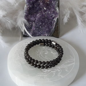 Op de afbeelding: Een dubbele armband van donkergrijze kralen met een zilveren metalen sluiting. De armband wordt getoond op een wit oppervlak met een decoratief ontwerp.