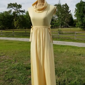 Pode incluir: Um vestido maxi amarelo claro com gola alta e um la&ccedil;o na cintura. O vestido &eacute; sem mangas e tem uma silhueta simples e fluida.