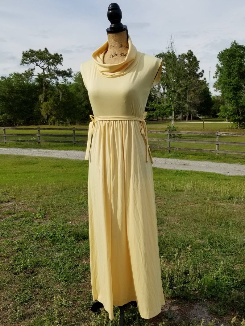 Pode incluir: Um vestido maxi amarelo claro com gola alta e um la&ccedil;o na cintura. O vestido &eacute; sem mangas e tem uma silhueta simples e fluida.