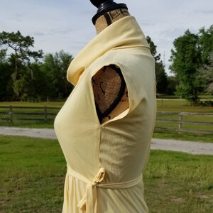 Pode incluir: Um vestido amarelo claro sem mangas com gola alta e cinto de n&oacute;. O vestido est&aacute; em um manequim contra um fundo de campo gramado e uma cerca de madeira.