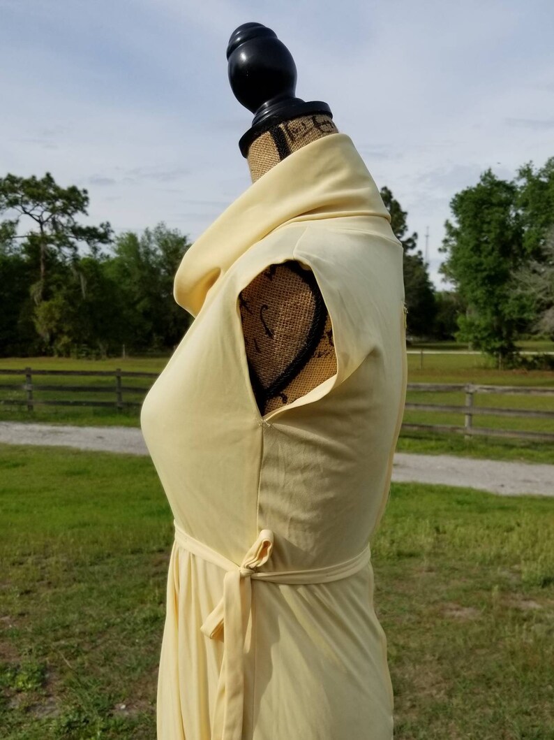 Pode incluir: Um vestido amarelo claro sem mangas com gola alta e cinto de n&oacute;. O vestido est&aacute; em um manequim contra um fundo de campo gramado e uma cerca de madeira.