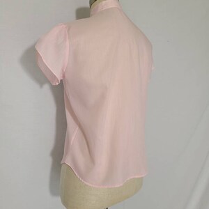 Pode incluir: Uma blusa rosa claro, transparente, com mangas curtas bufantes. A blusa tem um fecho de bot&otilde;es na frente e uma bainha arredondada.