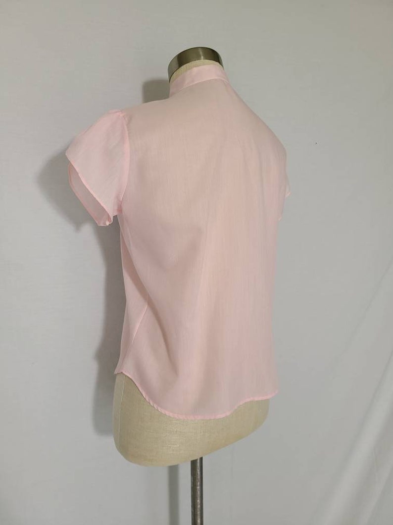 Pode incluir: Uma blusa rosa claro, transparente, com mangas curtas bufantes. A blusa tem um fecho de bot&otilde;es na frente e uma bainha arredondada.