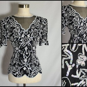 Puede incluir: Una blusa negra y blanca con lentejuelas, escote transparente y mangas cortas. La blusa tiene un cuerpo ajustado y un dobladillo festoneado.