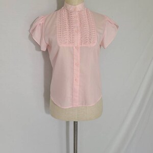 Pode incluir: Uma blusa rosa claro de manga curta com gola alta e fecho de bot&otilde;es na frente. A blusa tem um detalhe de babados decorativos na frente.