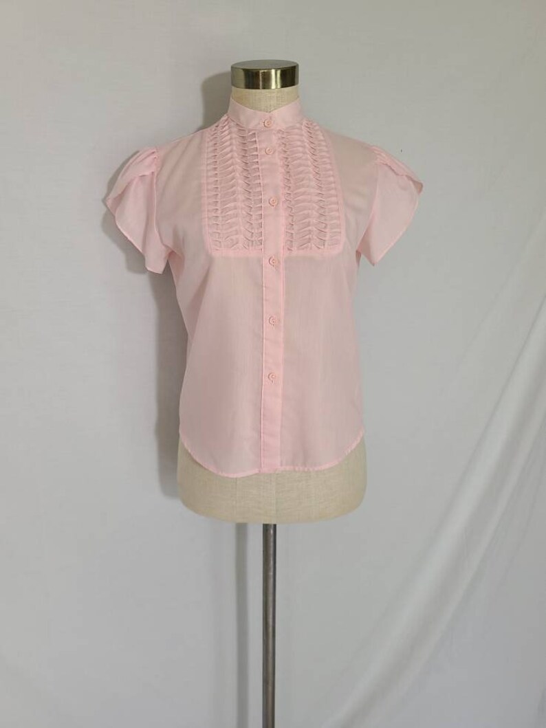 Pode incluir: Uma blusa rosa claro de manga curta com gola alta e fecho de bot&otilde;es na frente. A blusa tem um detalhe de babados decorativos na frente.