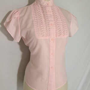 Pode incluir: Uma blusa rosa claro de manga curta com gola alta e uma frente plissada. A blusa tem fecho de bot&otilde;es.