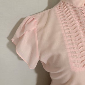 Pode incluir: Uma blusa rosa claro com colarinho alto e mangas curtas com babados. A blusa tem um detalhe franzido na frente.