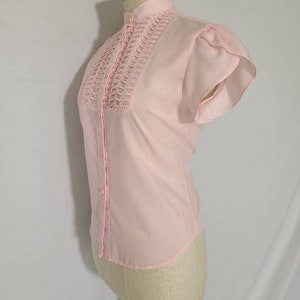 Pode incluir: Uma blusa rosa claro com gola alta e uma frente plissada. A blusa tem mangas curtas e bufantes.