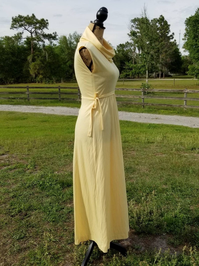 Pode incluir: Um vestido maxi amarelo claro sem mangas com gola alta e um la&ccedil;o na cintura.