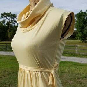 Pode incluir: Um vestido amarelo claro sem mangas com gola alta e um la&ccedil;o na cintura.