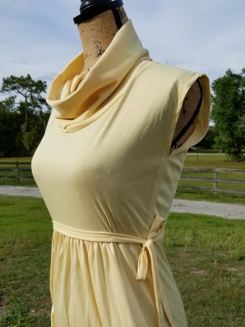 Pode incluir: Um vestido amarelo claro sem mangas com gola alta e um la&ccedil;o na cintura.