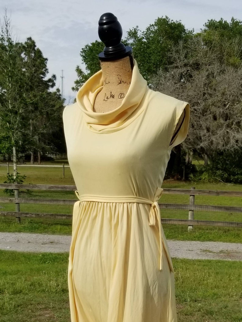 Pode incluir: Um vestido amarelo claro sem mangas com gola alta e um la&ccedil;o na cintura.