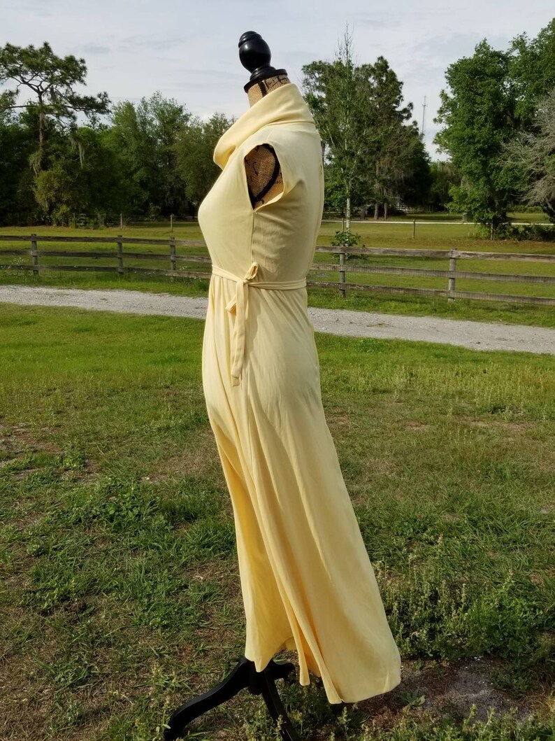 Pode incluir: Um vestido maxi amarelo claro com gola alta e mangas curtas. O vestido tem um la&ccedil;o na cintura e uma saia fluida.