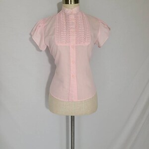 Pode incluir: Uma blusa rosa de bot&otilde;es com colarinho franzido e mangas curtas. A blusa est&aacute; em um manequim branco.
