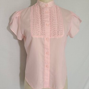Pode incluir: Uma blusa rosa claro de manga curta com gola alta e uma frente plissada. A blusa tem fecho de bot&otilde;es e uma bainha ligeiramente curva.