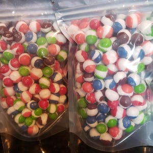 May include: Two clear plastic bags filled with colorful candies. The candies are small, round, and have a striped appearance with colors like red, white, blue, green, and brown. The bags are sealed with a zipper closure.