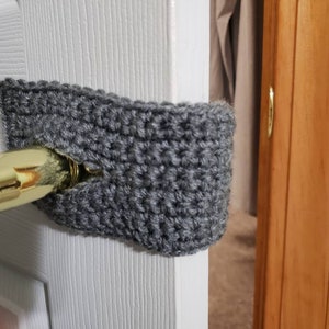 May include: A gray crocheted doorstop covering a gold door handle. The doorstop is made of a soft, textured yarn and is designed to prevent doors from slamming.