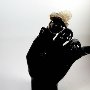 以下が含まれることがあります： 上に大きな白いクリスタルのような石が付いた金の指輪。指輪は黒い手につけられています。