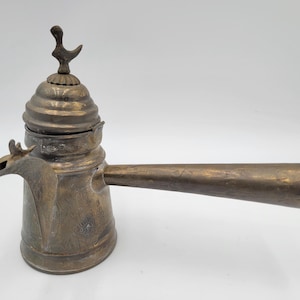 May include: Antique bronze-colored coffee pot with a long, horizontal handle. The pot has a decorative lid with a bird-like finial. The spout is shaped like a bird's head. The pot is likely made of brass or a similar metal.