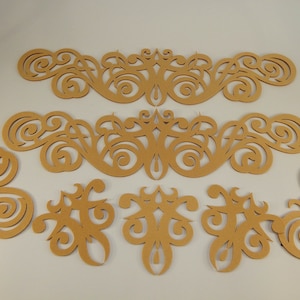 May include: Set of six laser-cut wood embellishments with intricate swirling patterns. The embellishments are in a light brown color and are arranged in a symmetrical pattern.