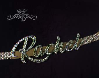 May include: The name "Rachel" is spelled out in rhinestones on a brown fabric strip. The rhinestones are iridescent and sparkle.
