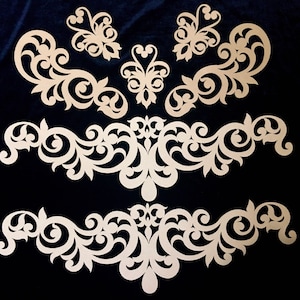 May include: Three decorative wooden cutouts with intricate floral designs. The cutouts are in the shape of a butterfly and two long, flowing designs.