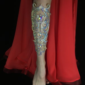 May include: A leg adorned with a decorative leg cuff made of rhinestones and gold-toned metal. The cuff features a floral design with a large, iridescent rhinestone in the center. The leg is wearing a red skirt.