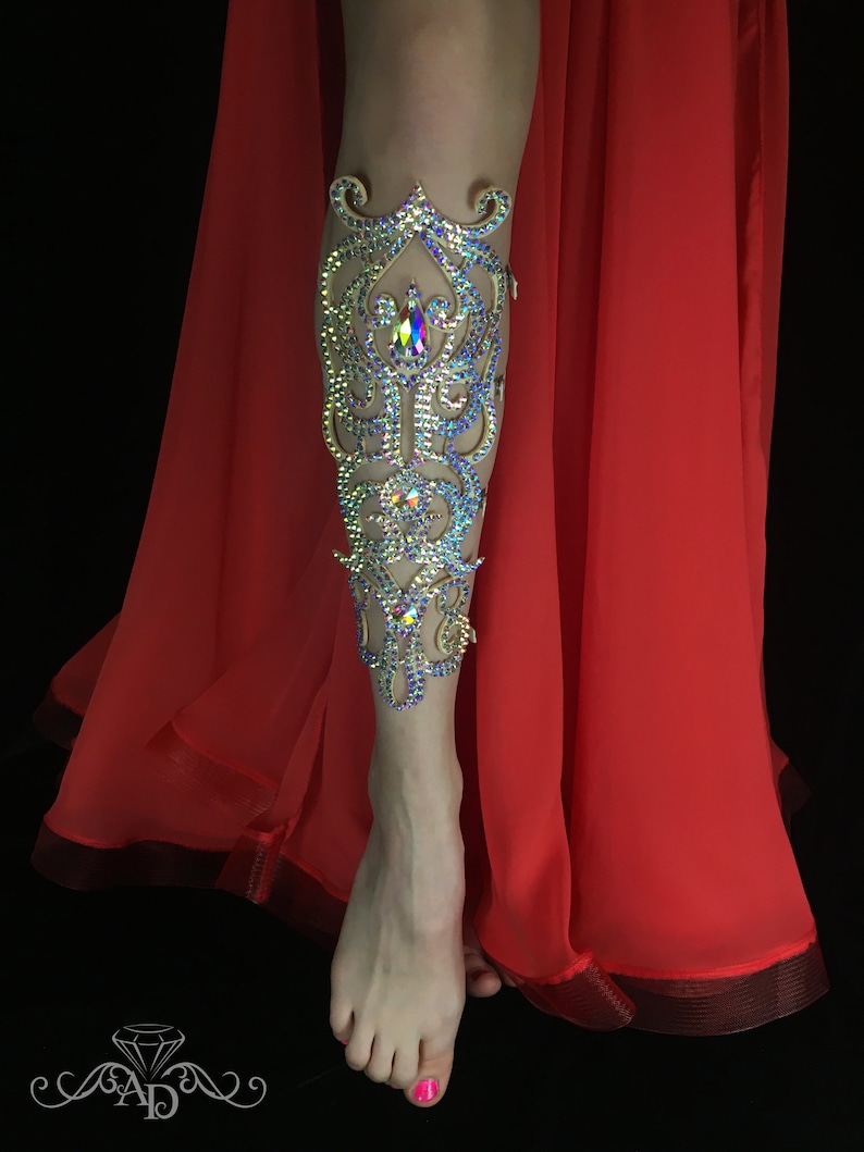 May include: A leg adorned with a decorative leg cuff made of rhinestones and gold-toned metal. The cuff features a floral design with a large, iridescent rhinestone in the center. The leg is wearing a red skirt.