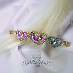 May include: A gold-toned bracelet with three heart-shaped charms made of iridescent rhinestones. The charms are in shades of pink, green, and blue.