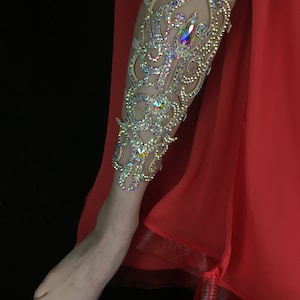 May include: A leg adorned with a decorative, ornate leg piece made of rhinestones and crystals. The leg piece is a light beige color and features a swirling, floral design. The leg is wearing a red skirt.