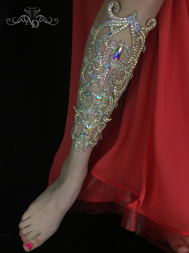 May include: A leg adorned with a decorative, ornate leg piece made of rhinestones and crystals. The leg piece is a light beige color and features a swirling, floral design. The leg is wearing a red skirt.