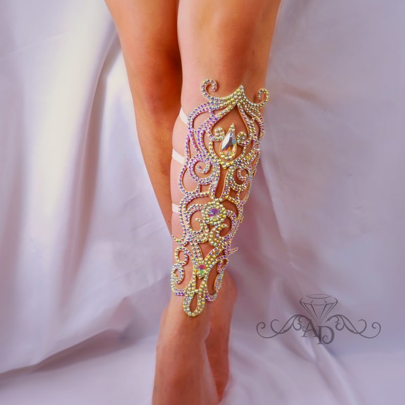 May include: A leg adorned with a decorative, ornate leg cuff. The cuff is made of white fabric and is covered in iridescent rhinestones. The cuff features a floral design with intricate swirls and curves.