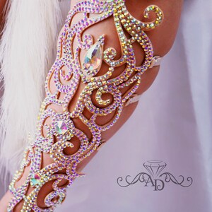 May include: A beige arm cuff with a decorative design made of iridescent rhinestones. The design features a floral pattern with a large teardrop-shaped rhinestone in the center.