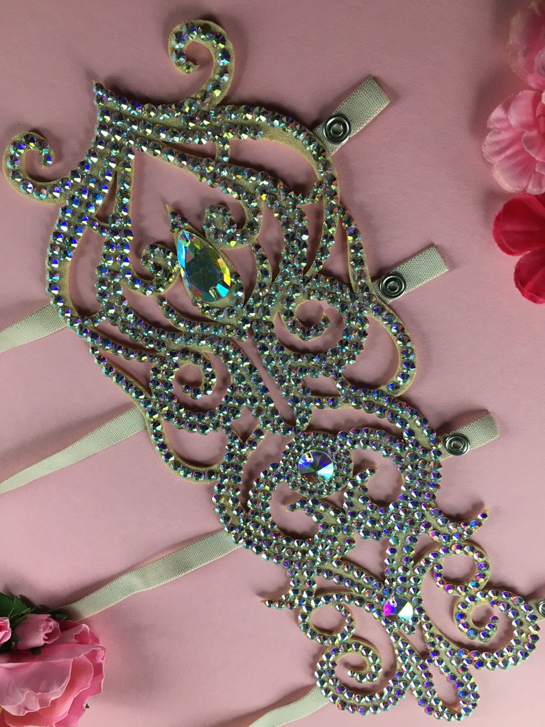 May include: A decorative embellishment for dance costumes, featuring a delicate, ornate design with iridescent rhinestones and a large crystal in the center. The embellishment is attached to a white ribbon with snaps.
