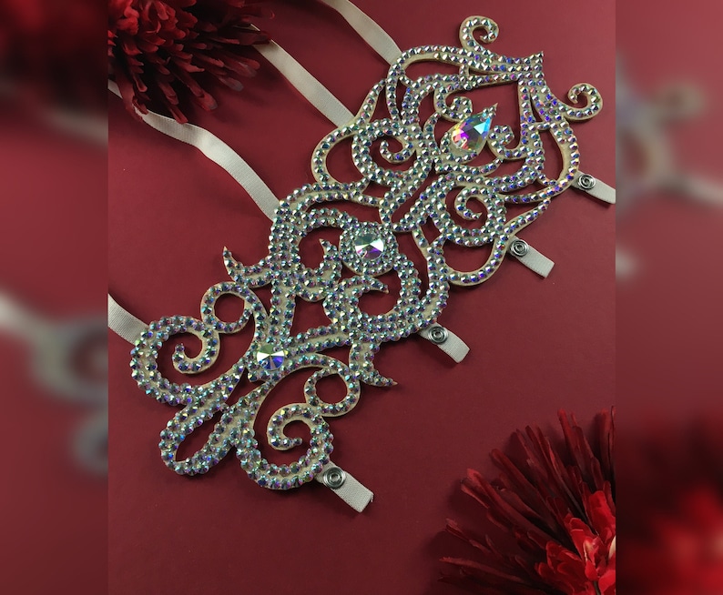 May include: A white, ornate, rhinestone-encrusted design with a floral pattern. The design is attached to white straps.