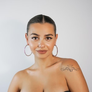 May include: A woman with dark hair is wearing large hoop earrings with iridescent rhinestones. She has a small tattoo on her shoulder that says "AD" with a diamond above the letters.