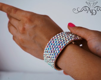 May include: A wide, sparkly, iridescent bracelet with a light brown leather base. The bracelet is covered in small, faceted rhinestones that create a shimmering effect.