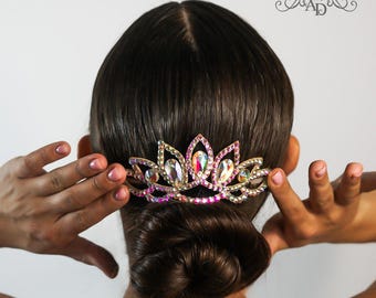 May include: A gold hair comb with iridescent crystals in a flower shape. The comb is being held in place by a person's hand.